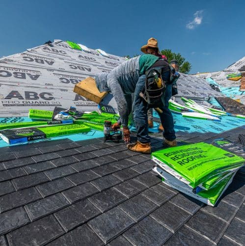 Roofer kneeling on a shingle roof inspecting damage while holding a clipboard