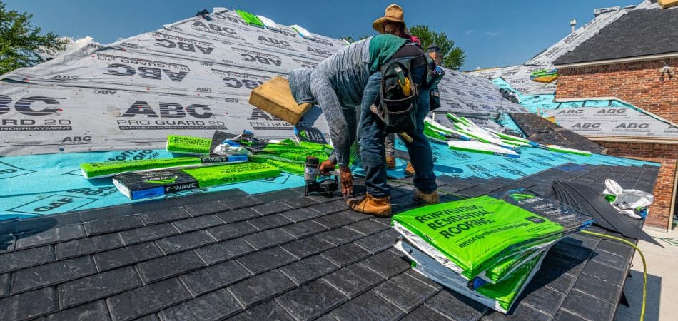 Roofer kneeling on a shingle roof inspecting damage while holding a clipboard